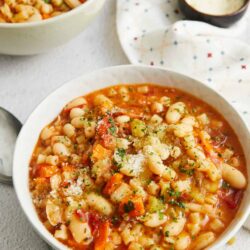 Creamy vegetable soup with white beans in a white bowl, garnished with herbs and cheese.