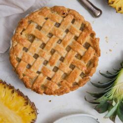 Golden pineapple pie with lattice crust, fresh pineapple slices, and baking utensils on a white surface.