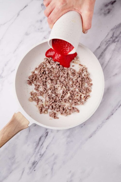 Minced meat being cooked in a skillet, with tomato sauce poured over it, on a marble countertop.
