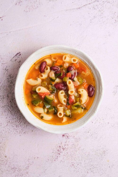 Creamy vegetable and pasta soup with beans in a white bowl on textured pink surface.