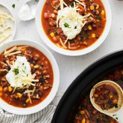 Savory beef chili in white bowls with shredded cheese and sour cream, side of corn, baked in slow cooker for effortless weeknight dinner.