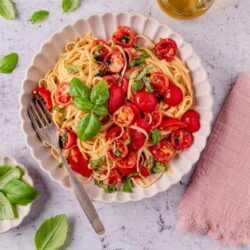 Basil and cherry tomato pasta served in a white scalloped bowl, fresh herbs, and a bottle of olive oil.