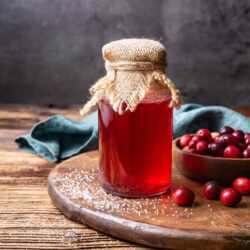 Fresh homemade cranberry jam in a jars with burlap cover, surrounded by cranberries on a wooden table.