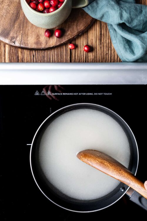 Fresh cranberries in a bowl on a wooden cutting board with a blue cloth, next to a sizzling milk pot on an induction stove.
