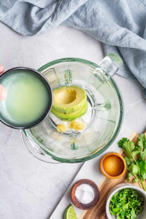 Creamy avocado and lime in a food processor with fresh herbs and seasonings ready for blending.