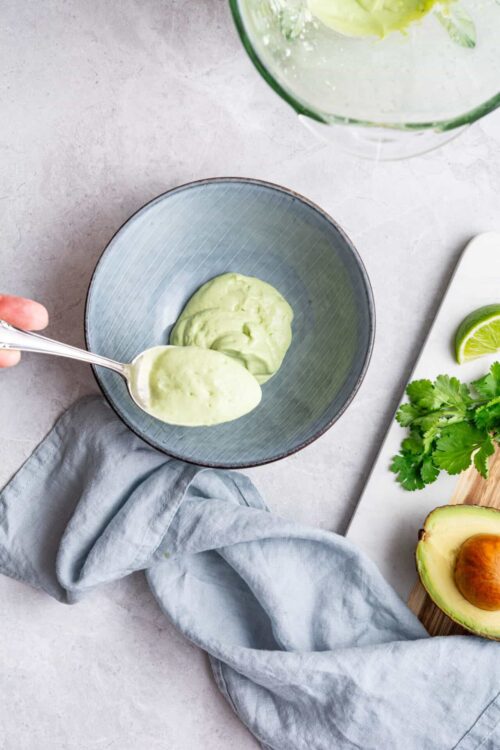 Creamy avocado sauce in a blue bowl on a light textured surface with fresh lime, cilantro, and half an avocado on a white cutting board.