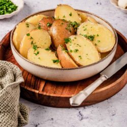 Golden roasted potatoes with fresh herbs in a white bowl on a wooden tray, ready to serve.