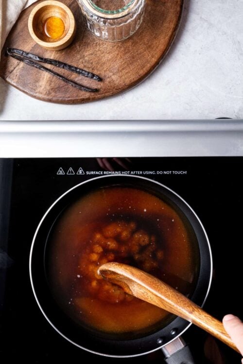 Vanilla extract in small bowl next to glass jar on wooden tray, close-up of caramel sauce being stirred in skillet, Baked Bree cooking blog image, homemade caramel recipe.