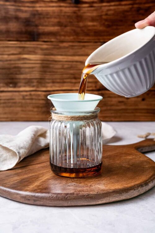 Light brown syrup being poured through a white cone into a glass jar, on a wooden cutting board.