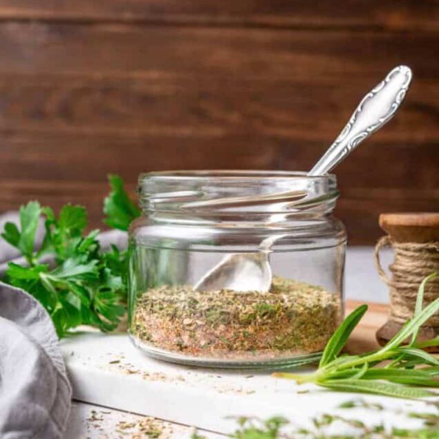 Dried herb blend in glass jar with spoon on rustic white surface, fresh herbs around, kitchen setting.