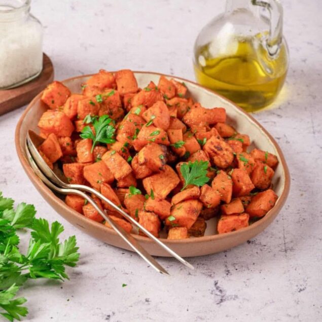 Crispy roasted sweet potato cubes garnished with fresh herbs on a beige platter.