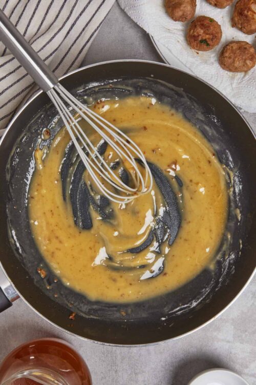 Creamy mustard sauce being whisked in a skillet with meatball ingredients in the background.