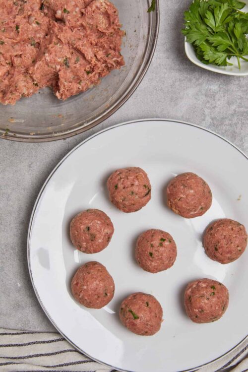 Seasoned raw meatballs with herbs on a white plate, ready for cooking.