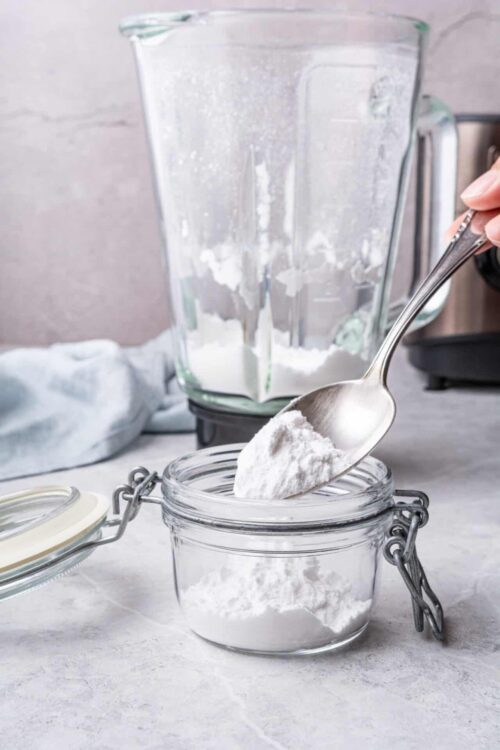 Fine white flour being measured with a spoon into a glass jar, with a blender in the background.