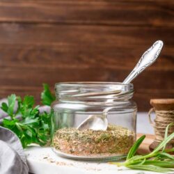 Dried herb spice blend in glass jar with fresh herbs and wooden spool, on white wooden surface.