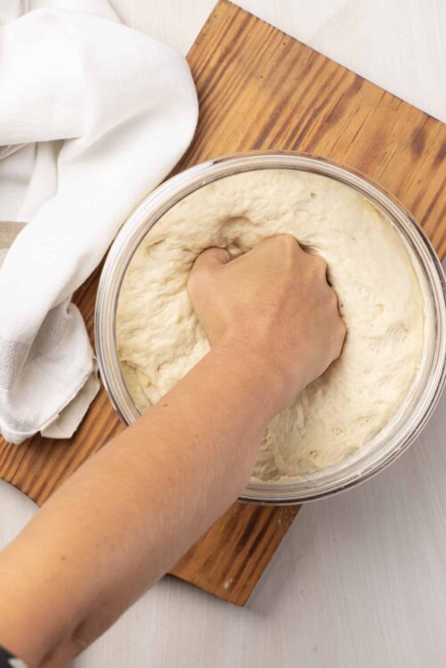 Fluffy homemade bread dough being kneaded in a glass bowl on a wooden surface.