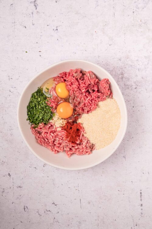 Ground beef, breadcrumbs, eggs, fresh herbs, garlic, and spices in a white bowl for cooking.
