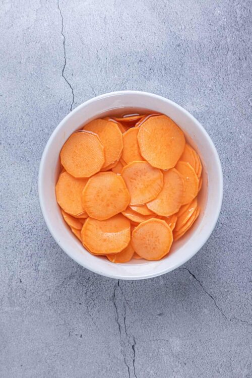 Sliced sweet potatoes in a white bowl on a concrete surface.
