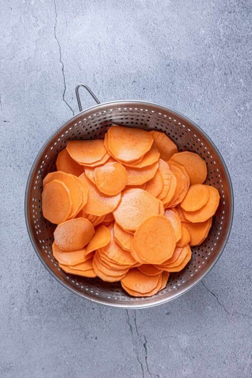 Sweet potato slices in a metal strainer on concrete surface.