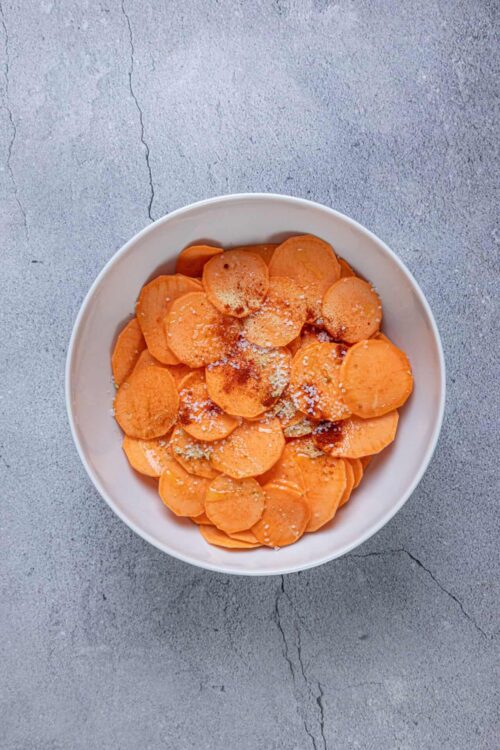 Sweet potato slices seasoned with spices in a white bowl on gray textured surface.