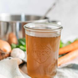 Golden homemade vegetable broth in a glass jar on a kitchen counter with fresh carrots, onions, and garlic.