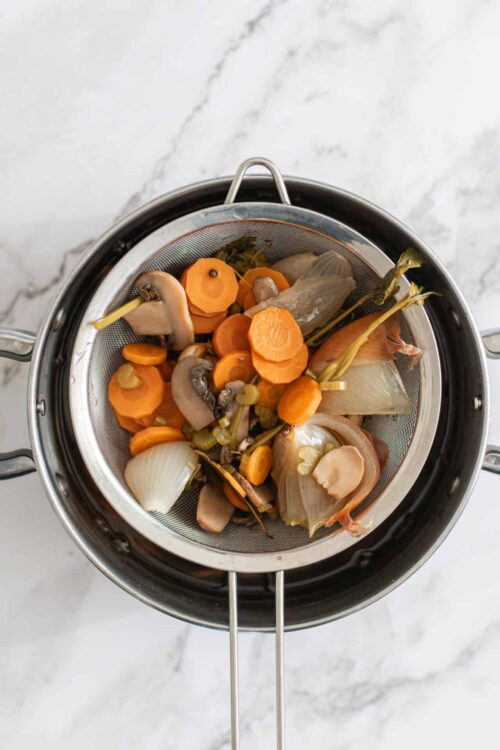 Steamed vegetable scraps in a fine mesh strainer over a pot, ready for flavoring or composting.