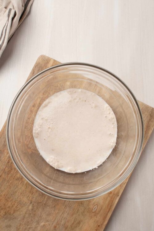 Creamy bread dough in glass bowl on wooden board, ready to bake bread or rolls.