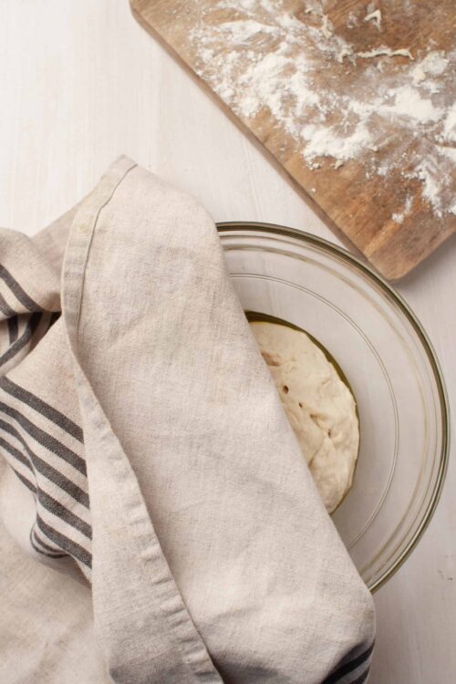 Soft cloth covering dough in glass bowl on white surface with floured wooden cutting board in background.