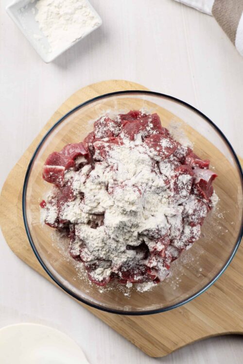 Chunks of raw beef with flour in a glass mixing bowl on wooden chopping board.