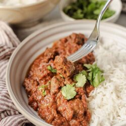 Savory beef stew over white rice in a beige bowl, garnished with cilantro, served with side dishes.
