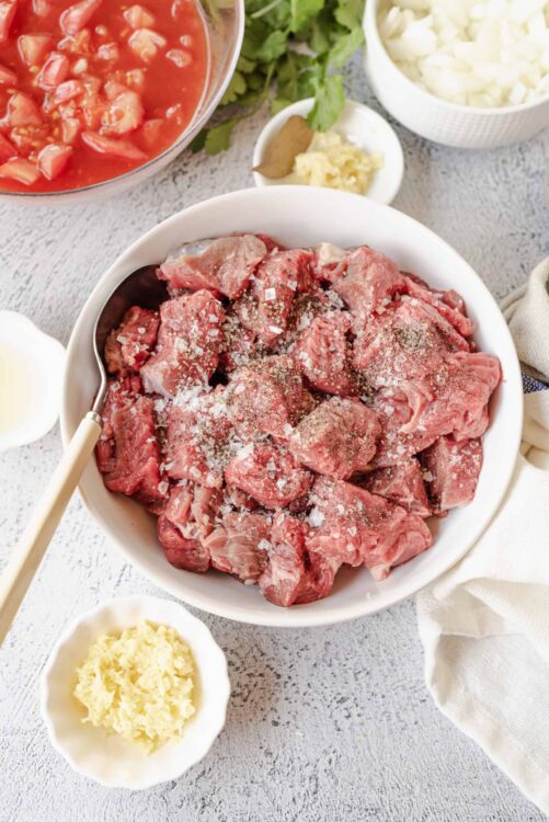 Chunks of raw beef with salt, pepper, and seasonings on a white plate for beef stew.
