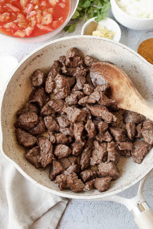 Braised beef stew in a skillet with cooking utensils and ingredients in the background, savory and hearty beef dish for comfort food recipes.