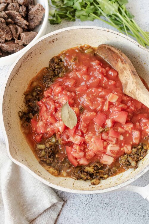 Savory beef and tomato stew cooking in a skillet with fresh herbs in the background.