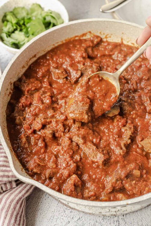 Savory beef chili stew in a beige pot with a spoon, topped with chopped fresh herbs.