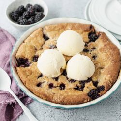 Juicy blackberry cobbler with vanilla ice cream served in a white ceramic dish on a textured gray surface.