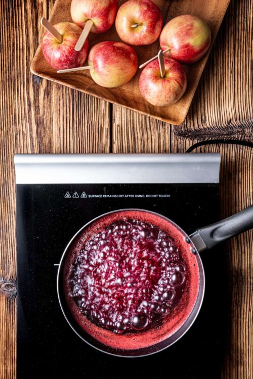 Fresh apples with skewers on a wooden tray, boiling berry sauce on a stovetop.