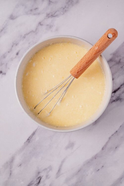 Smooth yellow batter in a white mixing bowl with a whisk and wooden handle, ready for baking.