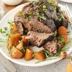 Juicy beef roast with roasted carrots and fresh herbs on white serving platter.
