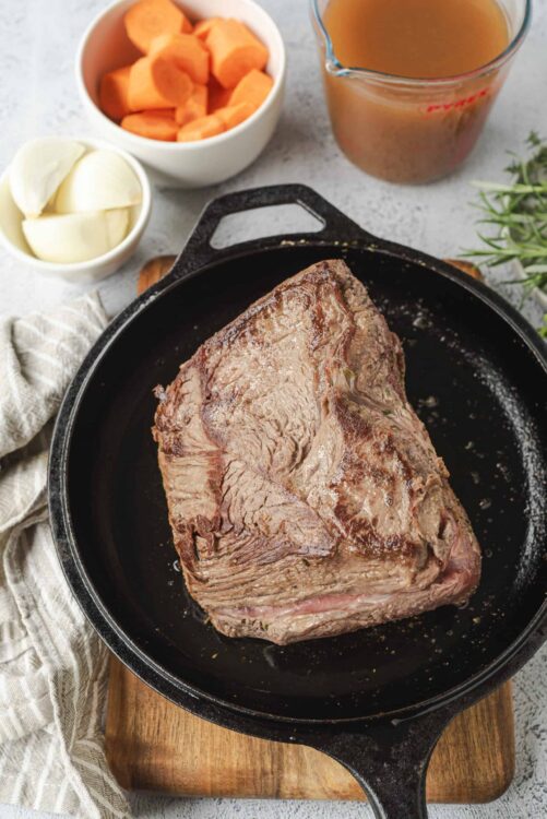 Juicy roast beef cooking in a cast iron skillet with carrots, onions, and fresh herbs.
