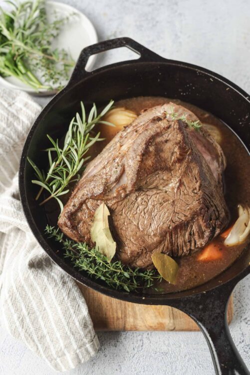 Juicy pot roast cooking in a cast iron skillet with fresh herbs, vegetables, and savory broth.