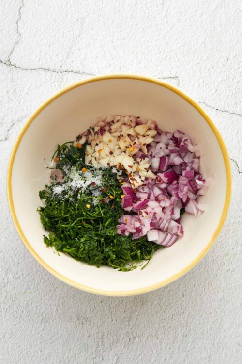 Diced red onions, garlic, chopped herbs, salt, and red pepper flakes in a bowl for cooking or baking.