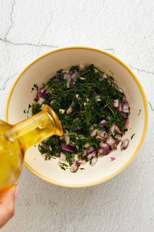 Minced red onions and herbs in a white bowl with olive oil being drizzled on top.