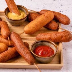 Crispy corn dog bites served with mustard and ketchup dips on a wooden tray.