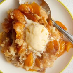 Sweet peach cobbler with vanilla ice cream on top, served in a white bowl. Perfect for dessert or summer treat.