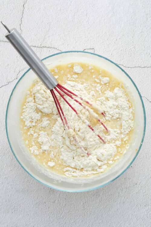 Cream mixture with flour and stand mixer paddle in a glass bowl on a textured white surface.