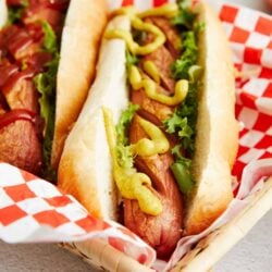 Grilled hot dog in bun with mustard, lettuce, and toppings in a basket.