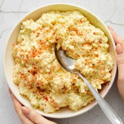 Creamy potato salad garnished with paprika in a large white bowl on a light background.
