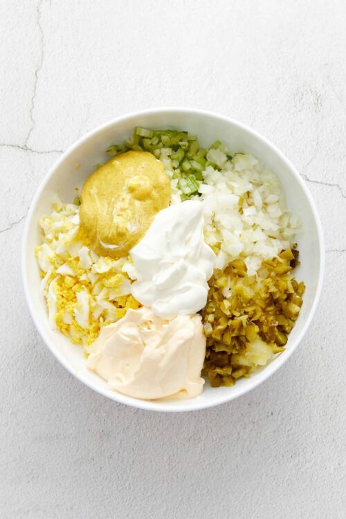 Creamy potato salad ingredients with mustard, mayonnaise, chopped celery, pickles, and hard-boiled eggs in a white bowl.