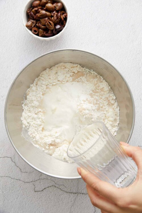 Crushed olives in a white bowl with flour and wet ingredients in a mixing bowl for baking.