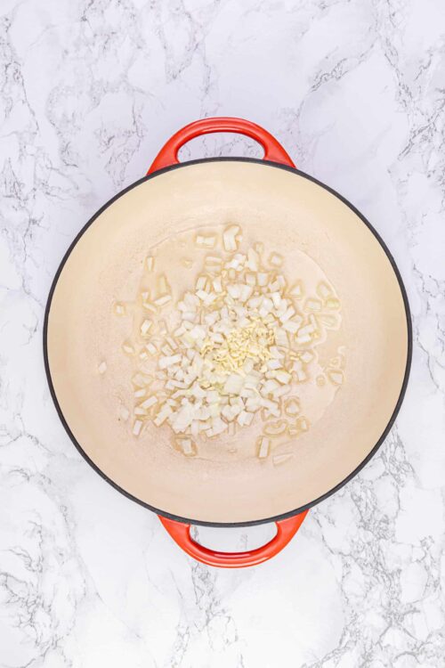 Diced onions and garlic sautéing in a cream-colored Dutch oven on a white marble countertop.